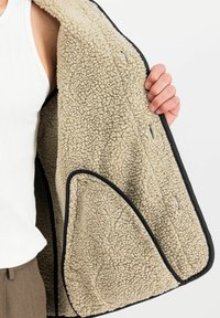 Veste en polaire beige avec un intérieur doux et texturé, bordée de finitions noires. Comprend des poches latérales et un design décontracté.