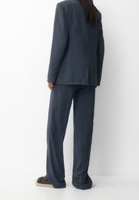 Completo su misura blu scuro con una vestibilità rilassata, caratterizzato da un blazer strutturato e pantaloni a gamba larga. Tessuto morbido e dettagli minimi.