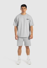 Camiseta de algodón gris con mangas cortas, combinada con pantalones cortos grises a juego. Presenta un detalle de logo en la camiseta y zapatillas deportivas blancas.