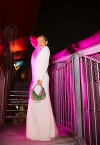 Lungo abito bianco fitted con maniche lunghe, abbinato a una borsa verde con tracolla a catena, su una scalinata illuminata da luci rosa.