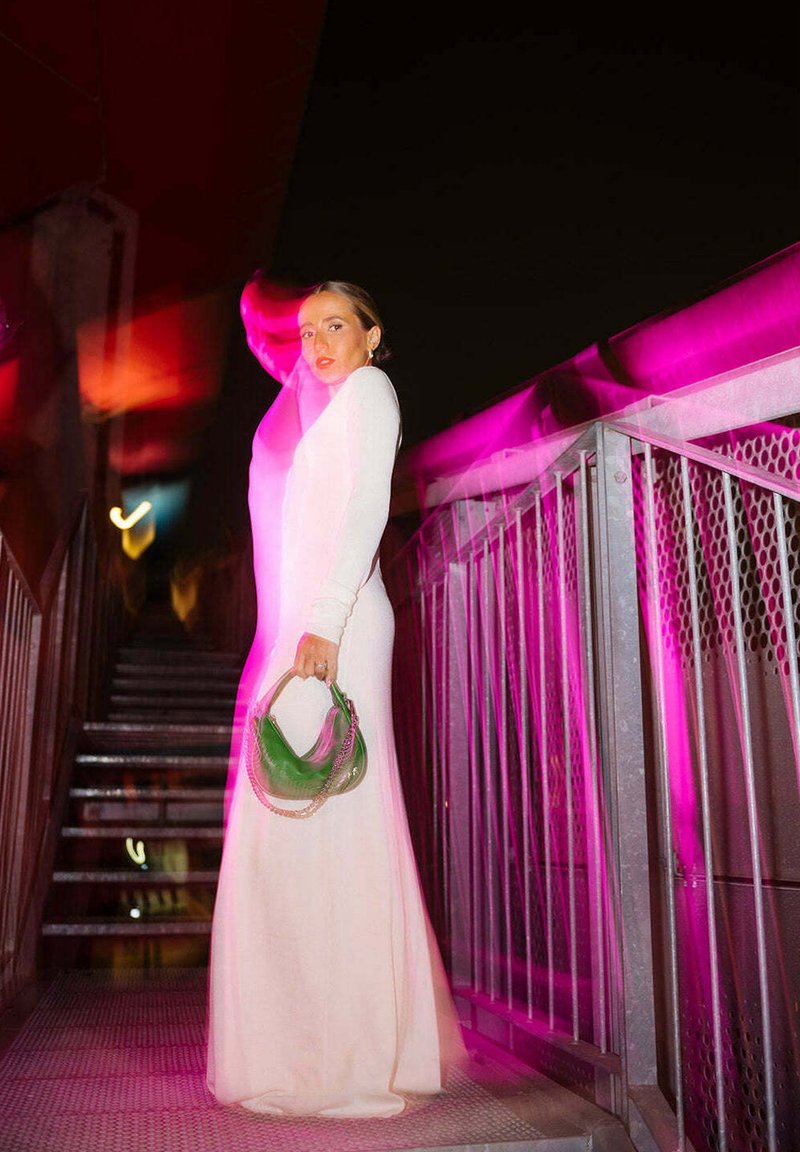 Lungo abito bianco fitted con maniche lunghe, abbinato a una borsa verde con tracolla a catena, su una scalinata illuminata da luci rosa.