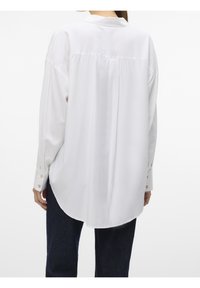Femme portant une chemise blanche ample à manches longues avec poignets boutonnés, vue de dos, associée à un pantalon foncé sur fond blanc.
