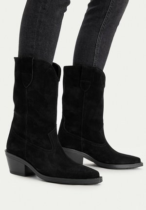 Black suede cowboy boots with mid-height block heels worn with dark fitted jeans against a white background.