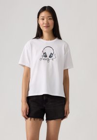 Νεαρή γυναίκα με μακριά μαλλιά, φοράει λευκό T-shirt με τυπωμένα μαύρα ακουστικά και τη λέξη "Levi's", συνδυασμένο με μαύρα τζιν σορτς.