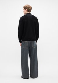 Μαύρος πουλόβερ με υφή στην πλάτη, συνδυασμένο με τζιν παντελόνι σε ίσια γραμμή και μπλε χρώμα με πίσω τσέπες. Το μοντέλο φαίνεται από πίσω.