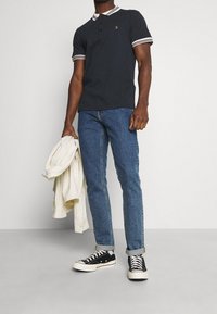 Dark navy polo shirt with white striped collar, paired with blue jeans and black sneakers, holding a cream shirt.