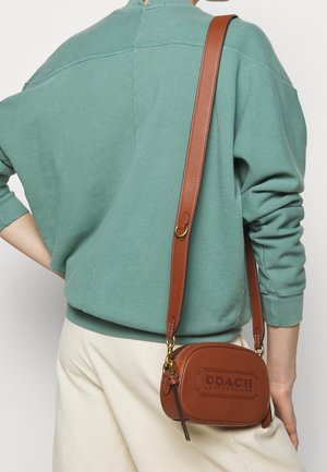 Bruine leren schoudertas met reliëflogo, rechthoekige vorm, gouden hardware en een verstelbare schouderriem, gedragen tegen een teal sweatshirt.