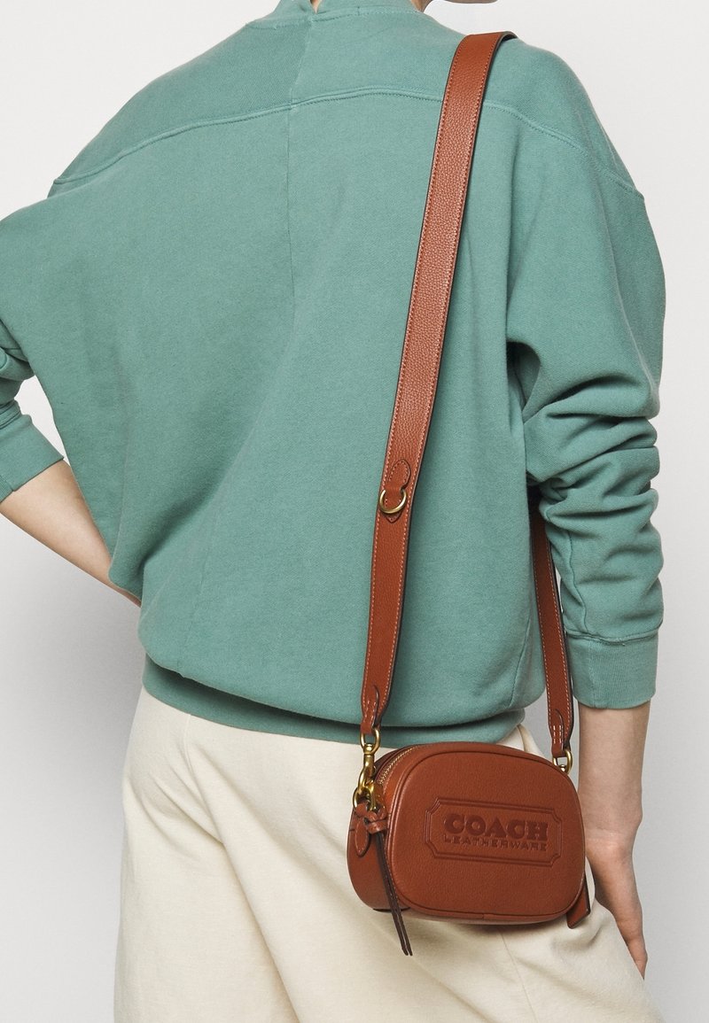 Brązowa skórzana torba crossbody z wytłoczonym logo, prostokątny kształt, złote akcesoria i regulowany pasek, noszona na tle turkusowego swetra.