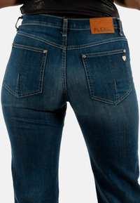 Jeans azules con una forma ajustada, fabricados en denim. Incluyen un parche de cuero marrón etiquetado "PLEASE" y acentos en hardware plateado.