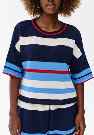 Kobieta w dzianinowym topie w paski - granatowym, białym, niebieskim i czerwonym oraz pasujących krótkich spodenkach, stojąca na białym tle.