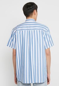 Camicetta a maniche corte in blu chiaro e bianco a righe verticali, realizzata in un tessuto dalla texture liscia. Presenta un colletto classico e una spalla posteriore.
