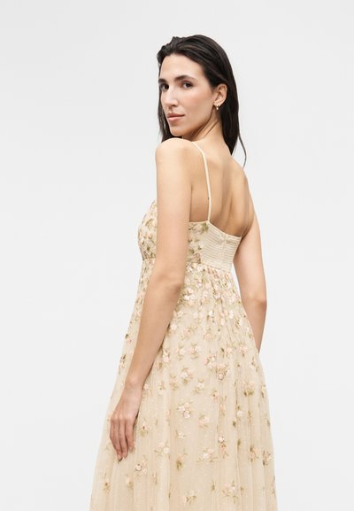 Γυναίκα με μακριά σκούρα μαλλιά, ντυμένη με αμάνικο μπεζ φόρεμα με κεντήματα με λουλούδια, κοιτάζει πάνω από τον ώμο της σε απλό φόντο.