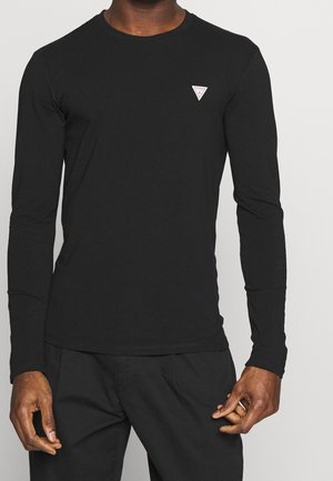 Mann trägt ein eng anliegendes schwarzes Langarmshirt mit kleinem weißem dreieckigem Logo auf der linken Brust, kombiniert mit schwarzen Hosen.