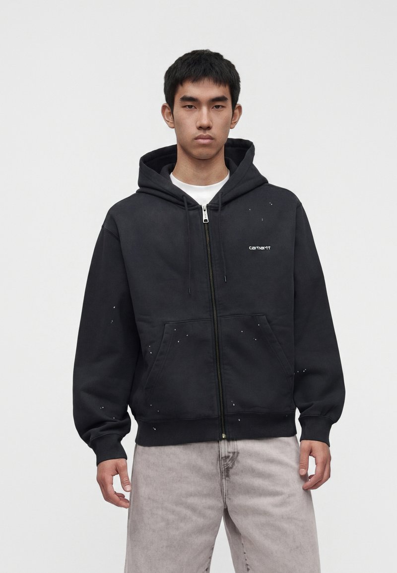 Jeune homme portant un sweat à capuche noir Carhartt zippé avec de petites éclaboussures de peinture blanche et un jean gris clair sur un fond uni.