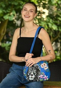 Blaue Handtasche mit einem Löwengesicht und floralen Designs. Hergestellt aus synthetischem Material mit glatter Textur und verstellbarem Tragegurt.