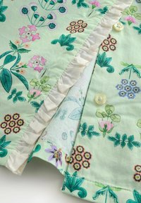 Vaaleanvihreä kangas monivärisillä kukkakuoseilla, valkoisella liehureunuksella ja pienillä kerma-nappeilla, joissa on kohokuvioitu teksti.