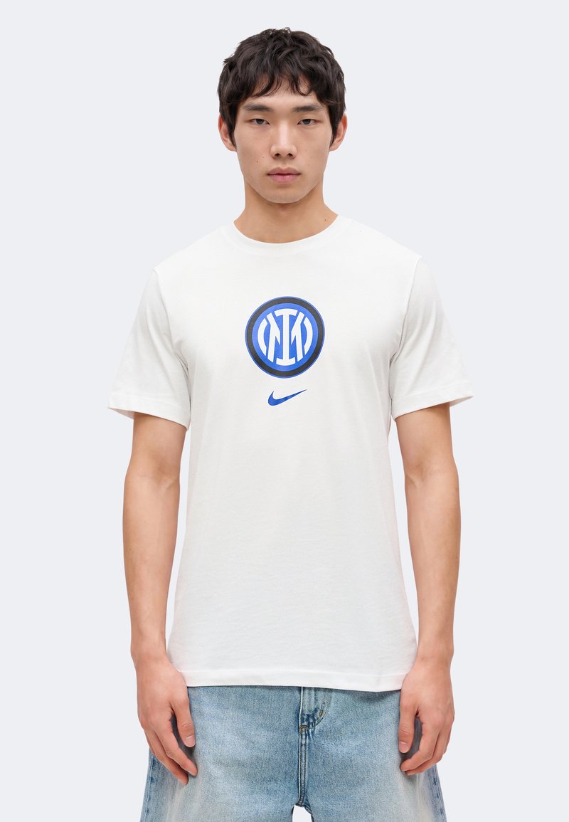 Jeune homme debout portant un t-shirt blanc avec un logo circulaire bleu et le swoosh Nike, associé à un jean en denim bleu clair.