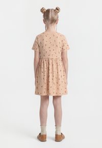 Szőke hajú gyermek két konttyal, barackszínű, apró virágmintás ruhában, krémszínű zokniban és barna cipőben, háttal állva.