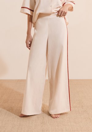 Pantalones crema de pierna ancha con una raya roja lateral, usados descalzos por una persona que está de pie sobre un suelo tejido.