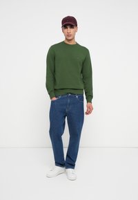 Egy férfi burgundi sapkát, zöld pulóvert, kék farmert és fehér sportcipőt visel, az egyik kezét zsebbe dugva áll egy fehér háttér előtt.