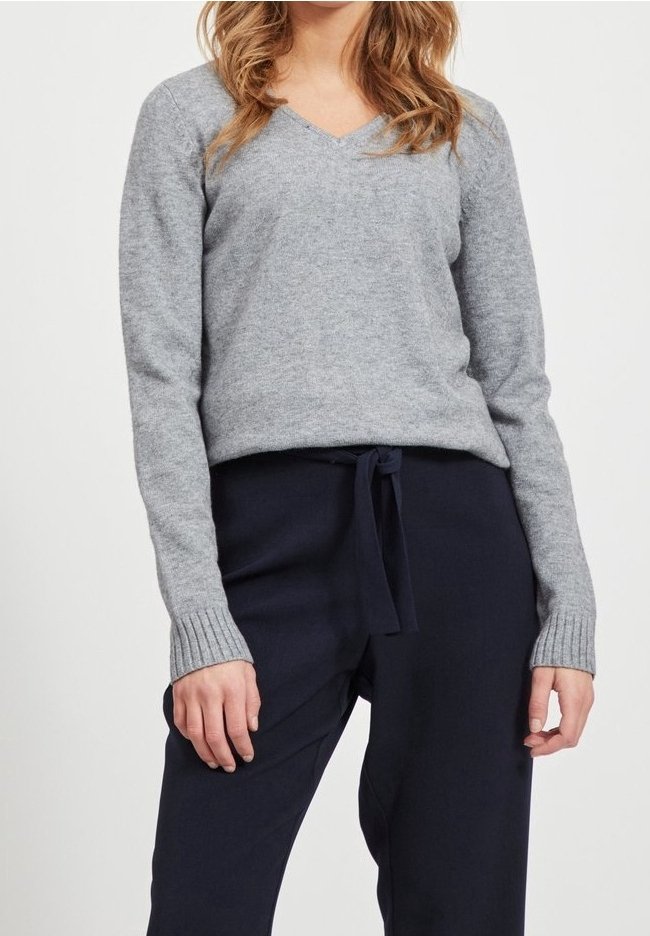 Femme portant un pull gris clair à col en V avec des poignets côtelés et un pantalon bleu marine foncé à cordon sur un fond uni.
