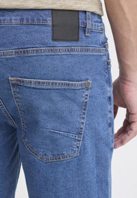 Siniset suoralahkeiset farkut, joissa takataskut ja musta nahkainen merkki vyötäröllä. Ompeleet näkyvissä.