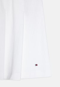 Hvit kortermet T-skjorte laget av bomull med en teksturert finish, med en liten brodert logo ved kanten i rødt, hvitt og blått.