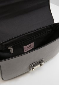 Šedá syntetická kabelka s elegantním designem, obsahující vnitřní zipovou kapsu a kovové zapínání. Podšívka s brandingovou nálepkou.