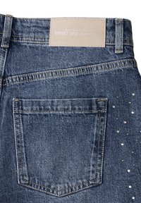 Ciemnoniebieskie dżinsy z tylną kieszenią, z beżową skórzaną łatą i porozrzucanymi srebrnymi ćwiekami wzdłuż boku dla ozdoby.