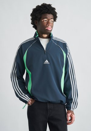 Tengerészkék cipzáras dzseki zöld részletekkel, fehér háromcsíkos ujjakkal és Adidas logóval. Könnyű anyag sima textúrával.
