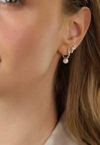 Pendientes de plata con un colgante de perla y un racimo de pequeñas piedras claras de un lado, con un diseño curvado y moderno.