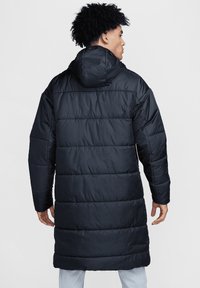 Langer, mit Kapuze versehener Puffermantel in Dunkelblau mit horizontalen Steppnähten. Glattes, glänzendes Material mit einem lässigen Schnitt, der bis zur Mitte der Oberschenkel reicht.