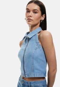 Top de mezclilla recortado en azul claro con cuello y cremallera frontal. Presenta un diseño sin mangas, costuras ajustadas y una silueta ceñida.