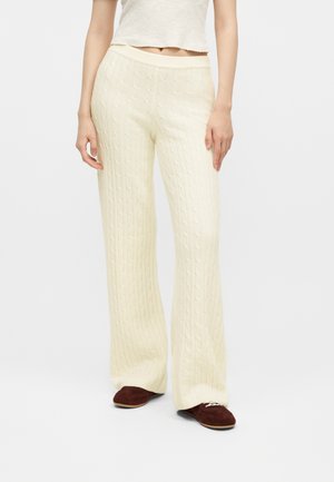 Femme portant un pantalon large en maille torsadée couleur crème et des chaussures marron à lacets, debout devant un fond uni.