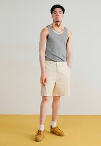 Grå tanktop, beige shorts, brune sko og hvite sokker. Lys hudfarge med synlige mønstre på armene. Enkle bakgrunn med gul gulv.
