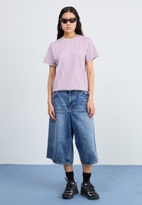Camiseta de color púrpura claro con escote redondo y logo. Culottes de denim azul con corte relajado, acompañados de zapatillas negras y calcetines.