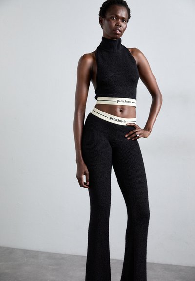 Woman wearing black sleeveless turtleneck crop top and matching high-waisted pants with white waistbands labeled "Palm Angels."
