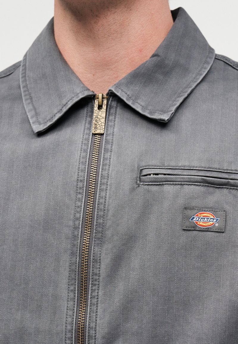 Primer plano del frente de una chaqueta gris Dickies que muestra cremallera metálica, cuello, bolsillo en el pecho y parche con el logo de la marca sobre tela con patrón espiga.