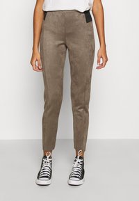 Pantalon en suède léger, de couleur marron clair, avec une coupe ajustée, doté de panneaux élastiques noirs à la taille, associé à des baskets montantes noires.