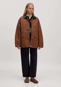Bruine oversized jas met een contrasterende zwarte kraag, voorzakken en drukknopen, gecombineerd met donkere broek en bruine pantoffels.