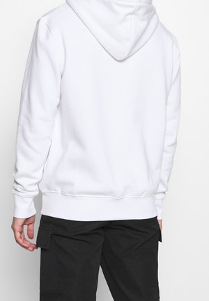 Person trägt einen schlichten weißen Hoodie und schwarze Cargo-Hosen, von hinten mit entspannter Haltung vor weißem Hintergrund gezeigt.