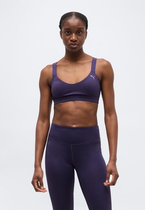 Femme athlétique portant un ensemble assorti de brassière de sport violette et de leggings taille haute, debout devant un fond clair uni.