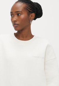 Λευκό φούτερ με λαιμόκοψη στρογγυλή και υφή στο ύφασμα. Διαθέτει διακριτικές κεντητές λεπτομέρειες στο στήθος. Το μοντέλο έχει πλεξούδες στα μαλλιά και σκουλαρίκια.