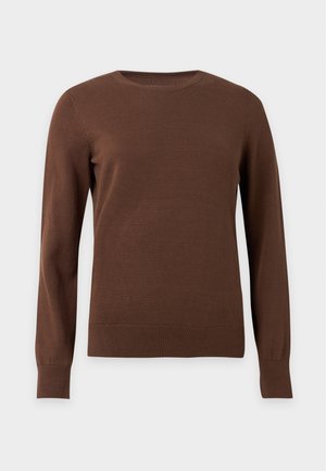 Femme aux cheveux courts et foncés avec des lunettes, portant un pull marron, un jean bleu à jambes larges et des bottes noires, debout avec une main dans la poche.