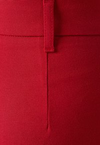 Rode stof close-up met een gladde textuur en subtiele weving, met een zichtbare naad en een klein verticaal tabje bij de taille.