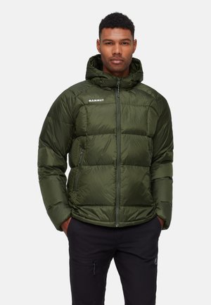 Mann trägt grüne Mammut Daunenjacke mit Kapuze und schwarze Hose, steht mit Händen in den Taschen vor einfarbigem Hintergrund.