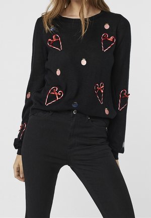 Schwarzer Pullover mit roten Ribbons in Herzform und kleinen rosa und blauen Verzierungen, getragen mit schwarzer Hose.