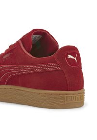 Zapatilla de ante rojo con suela de goma, que presenta costuras blancas, parche de logo característico y diseño de punta redondeada.