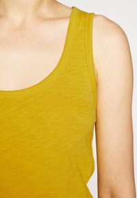 Lähikuva henkilöstä, joka käyttää hihattomaa sinapinkeltaista toppia, joka paljastaa olkapään, yläkäden ja solisluun alueen.