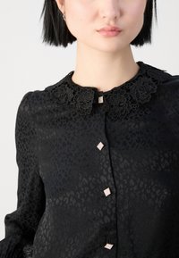 Blouse noire avec un imprimé léopard texturé. Un col en dentelle florale et des boutons décoratifs en forme de diamant mettent en valeur le devant. Manches longues.